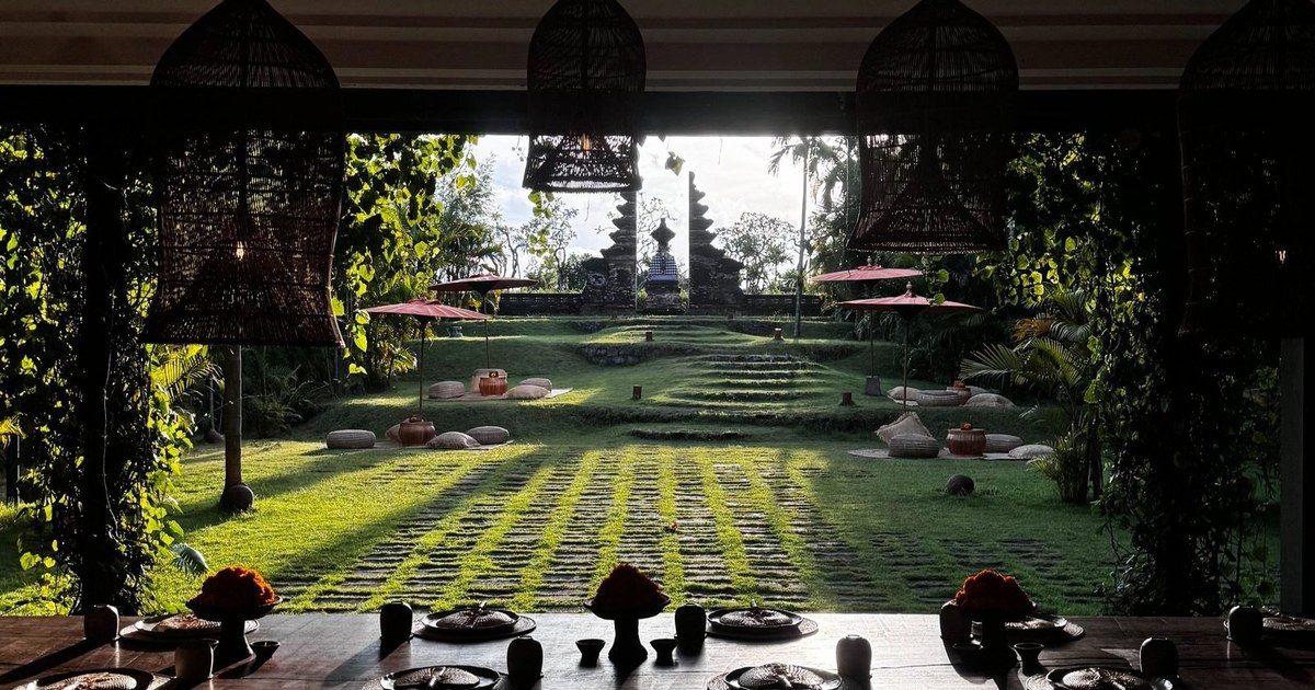 A long dining table under rattan lanterns looking across sunlit lawns to a Balinese temple gate