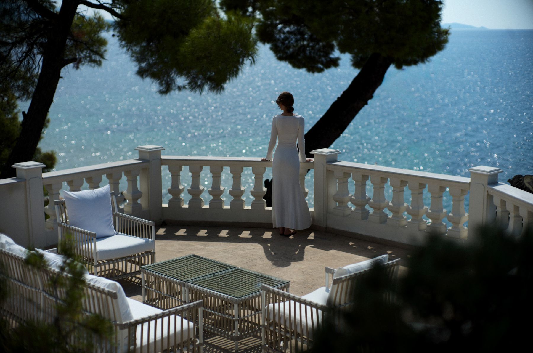 A guest in a long white dress at a white balustrade above the Aegean, beneath pines, at The Danai, Halkidiki