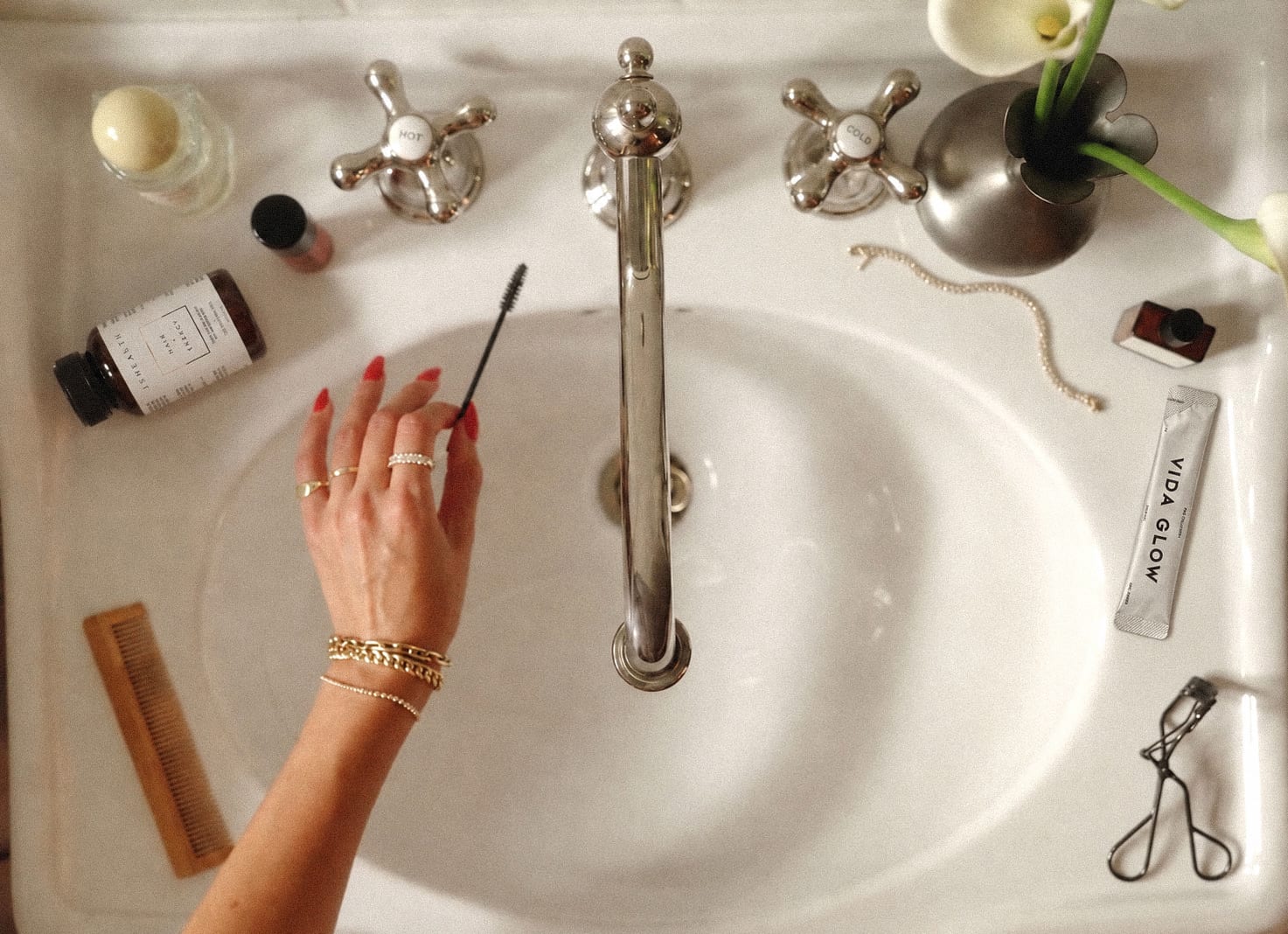 An overhead view of a hotel basin with chrome taps, a hand holding a lash wand, a JSHealth supplement bottle, a Vida Glow sachet and calla lilies
